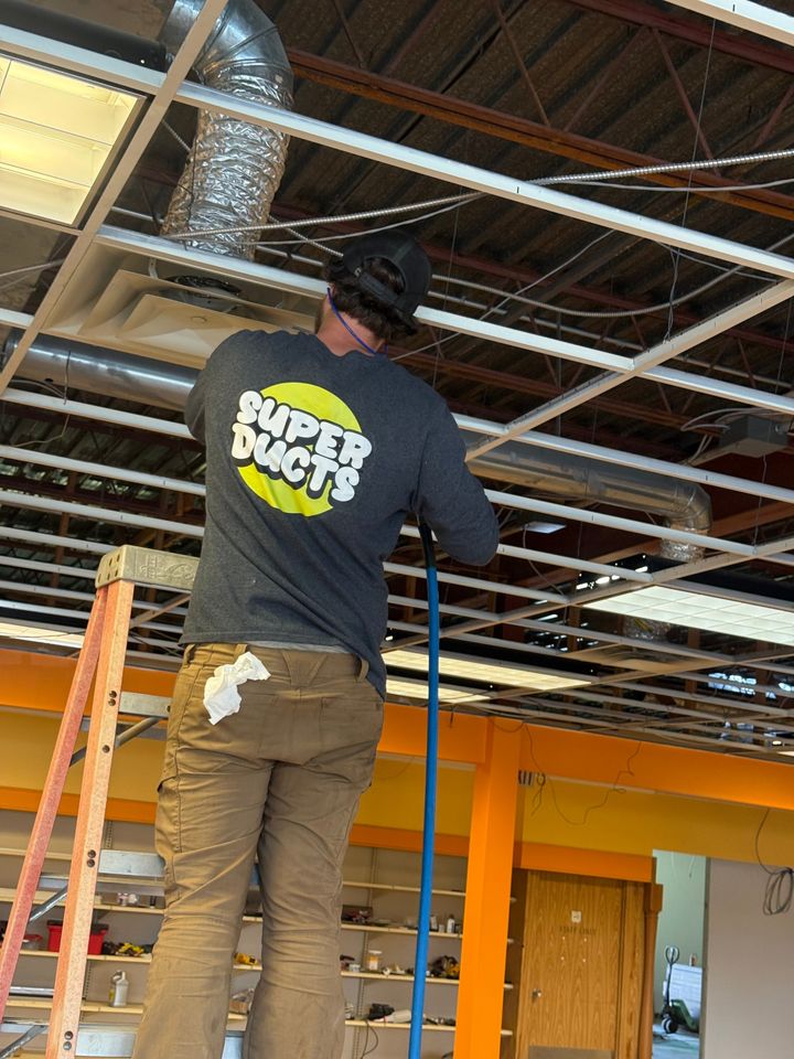 Super Ducts technician on a ladder holding a blue hose up to the ceiling ducts