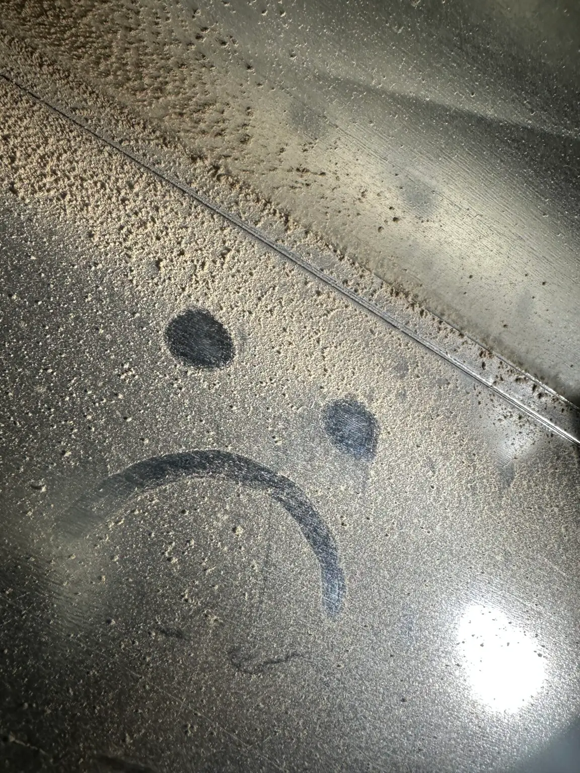A sad face is drawn with a finger in the heavy dust covering a reflective surface, highlighting the need for duct sealing to improve indoor air quality—especially important in Muskoka homes. Light reflects off the glass in the lower right corner.