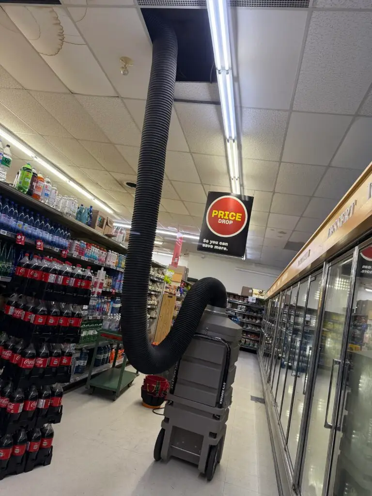 A large black hose extends from a ceiling vent to an industrial machine on wheels in a supermarket aisle, suggesting commercial duct cleaning Muskoka, surrounded by shelves of drinks and fridges. A Price Drop sign hangs nearby.